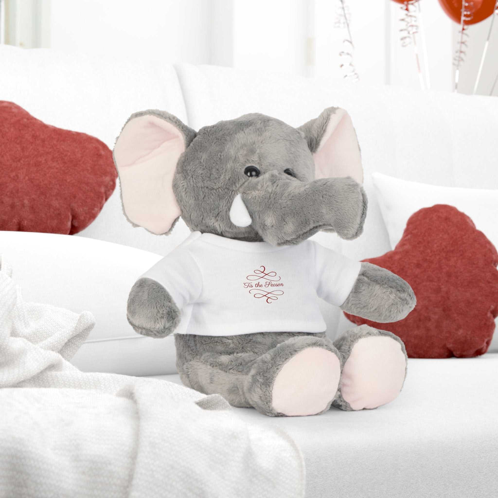 Gray plush elephant toy wearing white "Tis the Season" holiday t-shirt on couch with red pillows