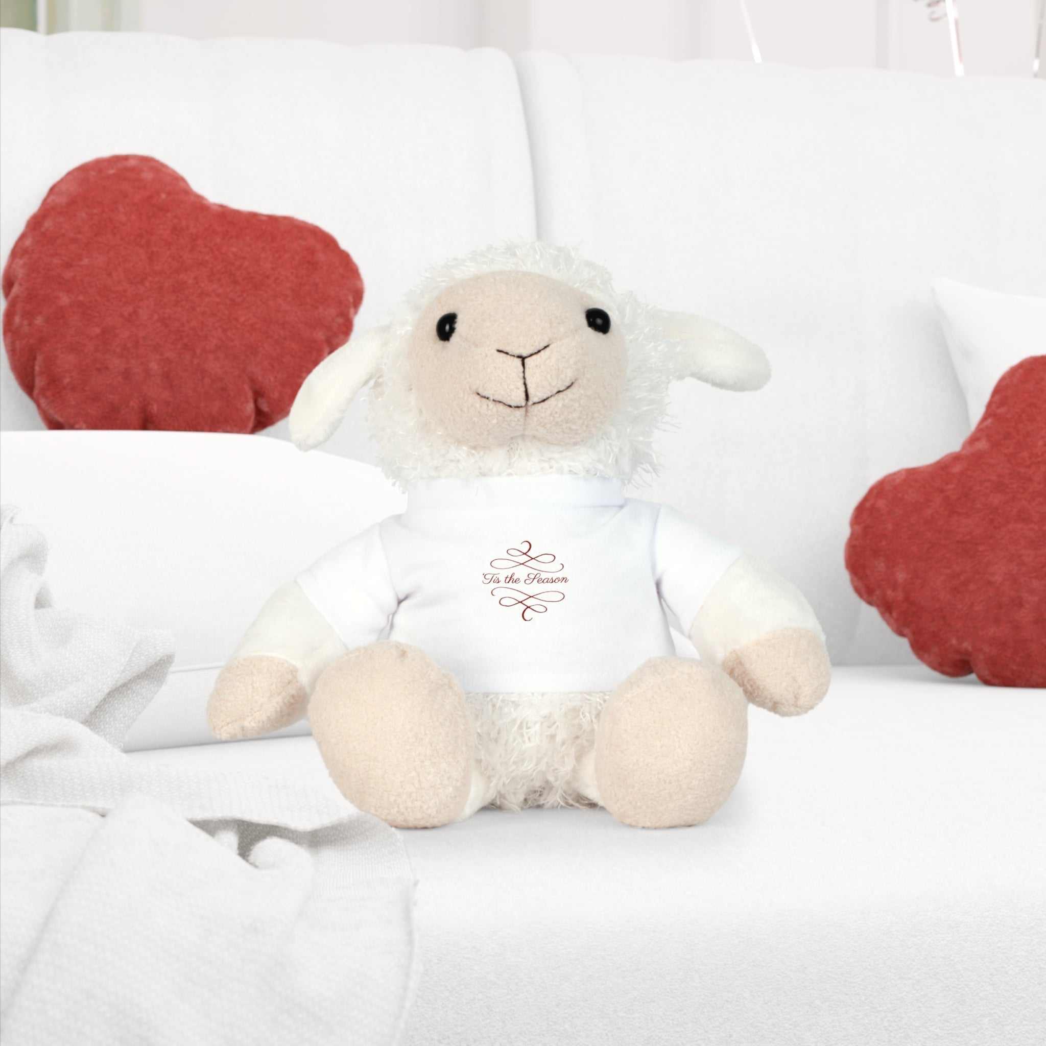 Soft white sheep plush toy wearing a white holiday t-shirt with "Tis the Season" text, sitting on a white couch with red cushions.
